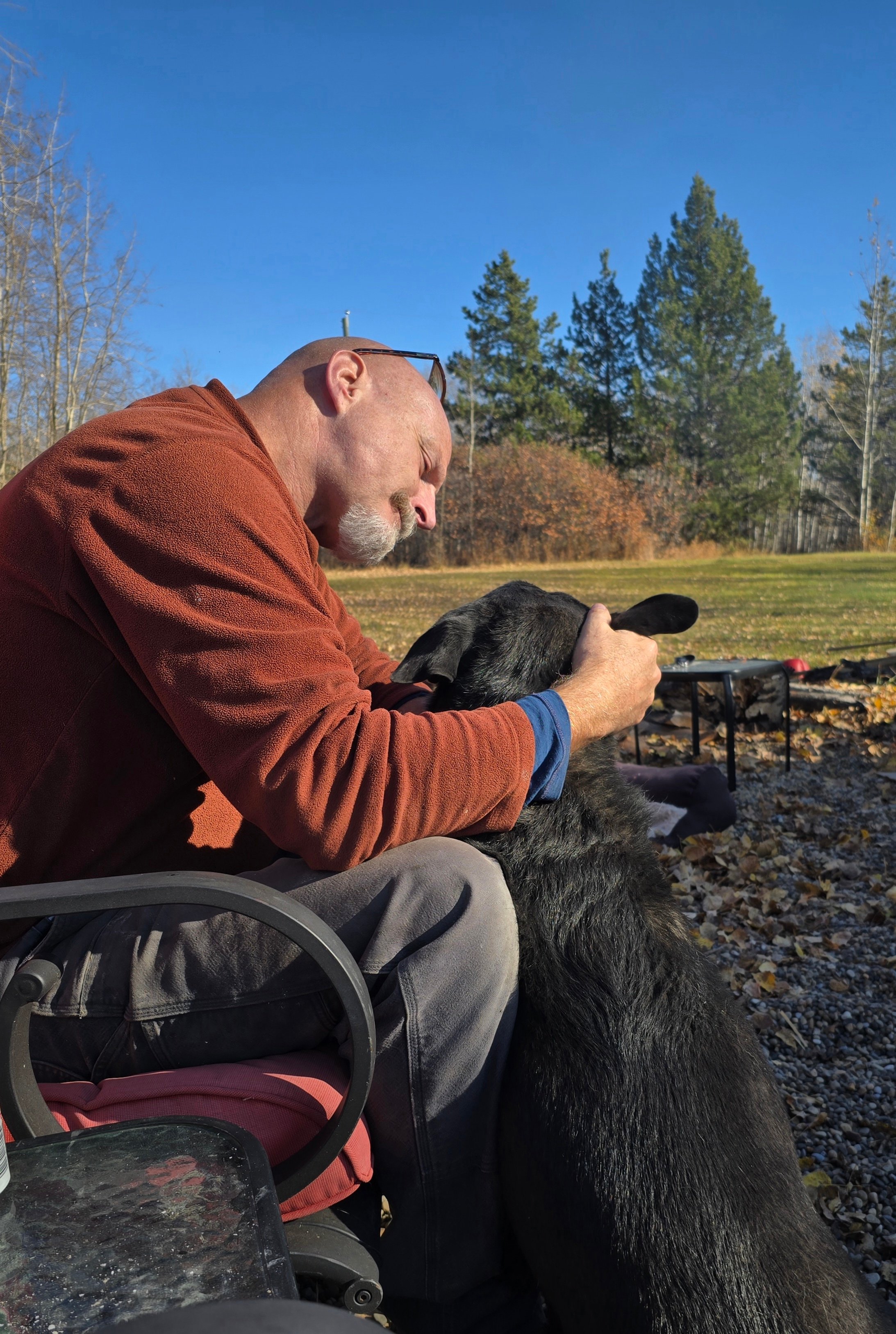 Man wearing rust jacket sitting outdoors petting a black dog on fall day with trees in background.